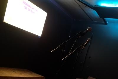 Deux micros sur pied dans une salle sombre, éclairée par un écran lumineux affichant un texte flou.