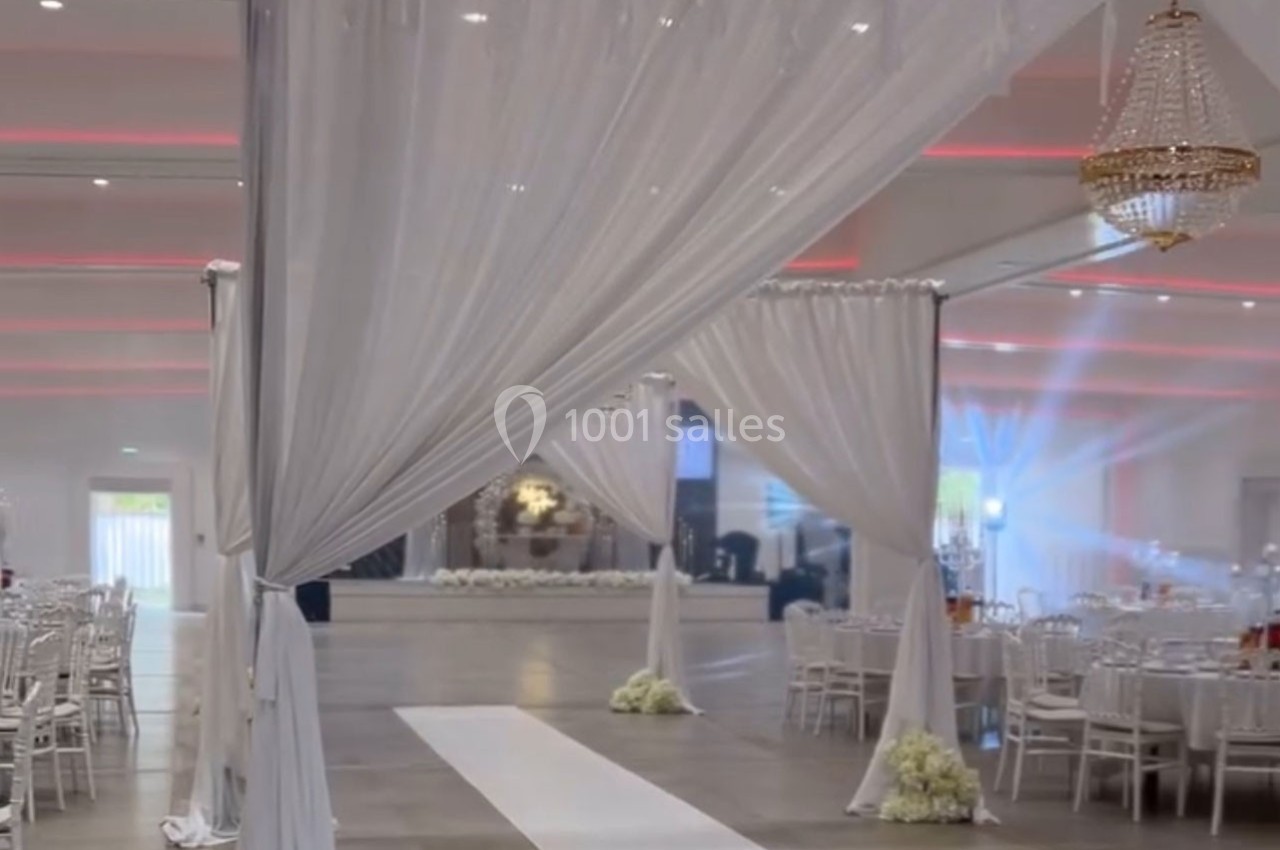 Salle de réception décorée avec des drapés blancs, un lustre, des tables dressées et une allée centrale.