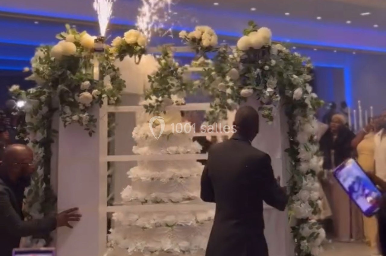 Un gâteau de mariage à plusieurs étages décoré de fleurs blanches, entouré d'arcs fleuris et de feux d'artifice.