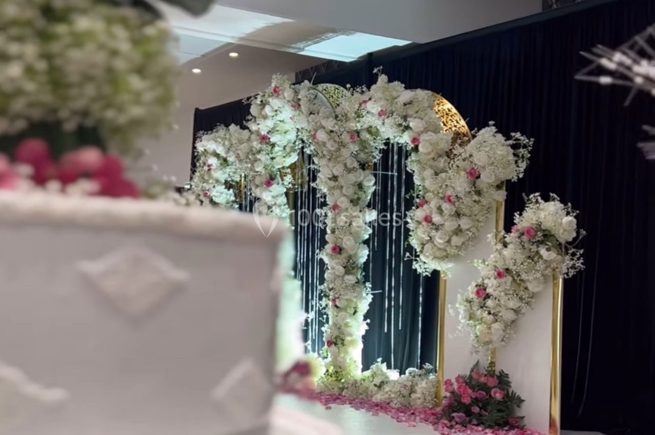 Arche décorative ornée de fleurs blanches et roses, avec un rideau noir en arrière-plan et un gâteau flou au premier plan.
