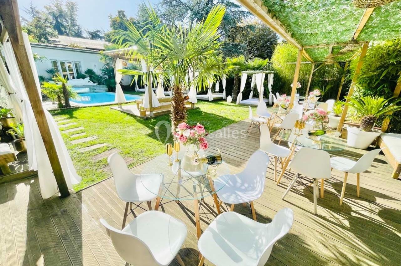 Terrasse en bois avec tables et chaises blanches, vue sur un jardin verdoyant, une piscine et des voiles décoratives…