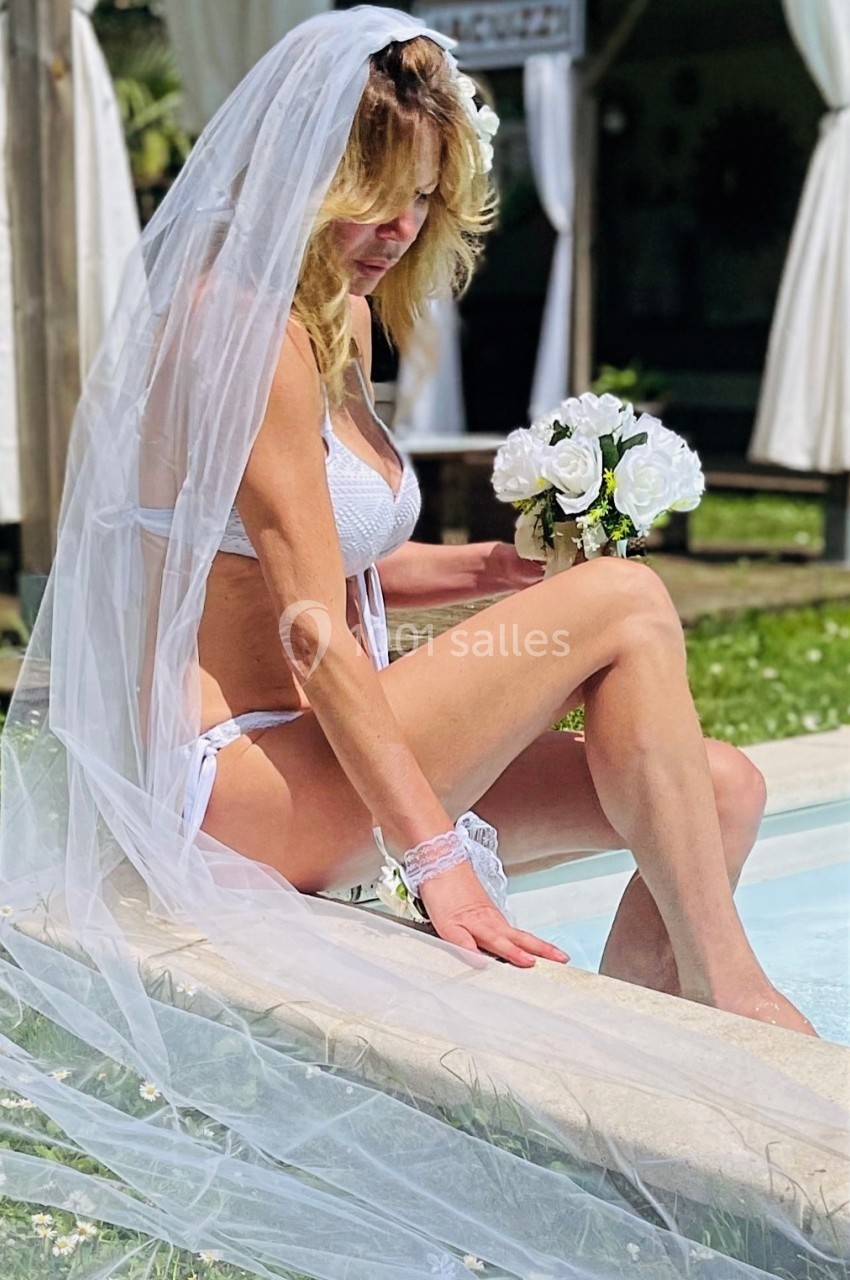 Femme en maillot de bain blanc et voile de mariée, assise au bord d'une piscine, tenant un bouquet de fleurs blanches.