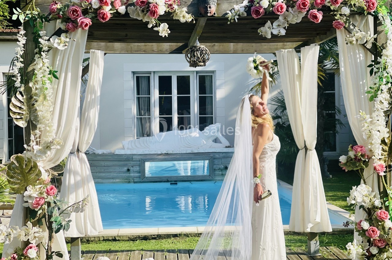 Une mariée en robe blanche lève son bouquet sous une pergola décorée de fleurs, près d'une piscine et d'une statue.