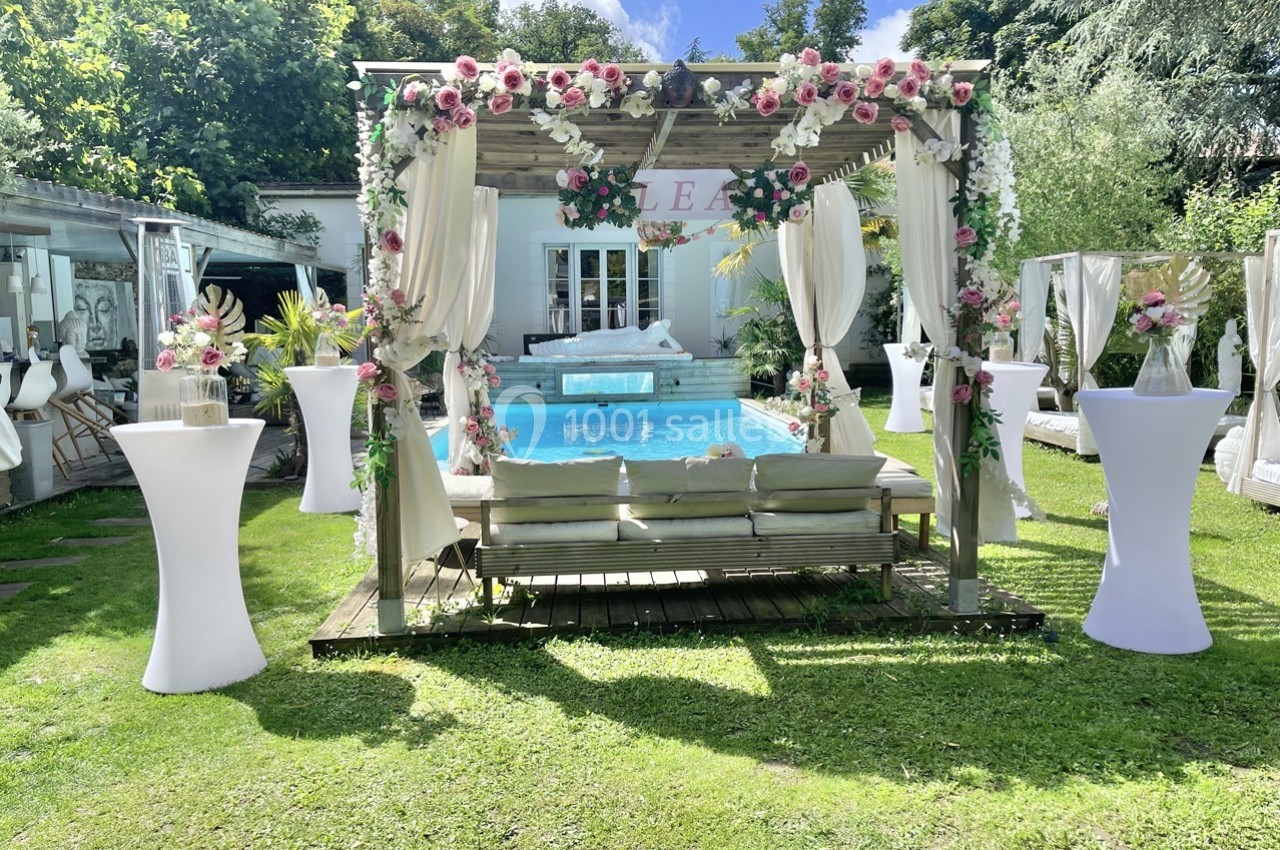 Pergola décorée de fleurs devant une piscine, entourée de verdure et de mobilier de réception en plein air.