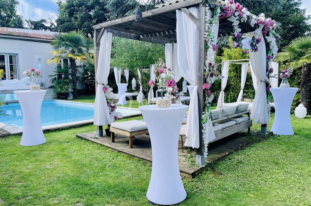 Pergola décorée de fleurs avec des tables hautes blanches, installée près d'une piscine dans un jardin verdoyant.