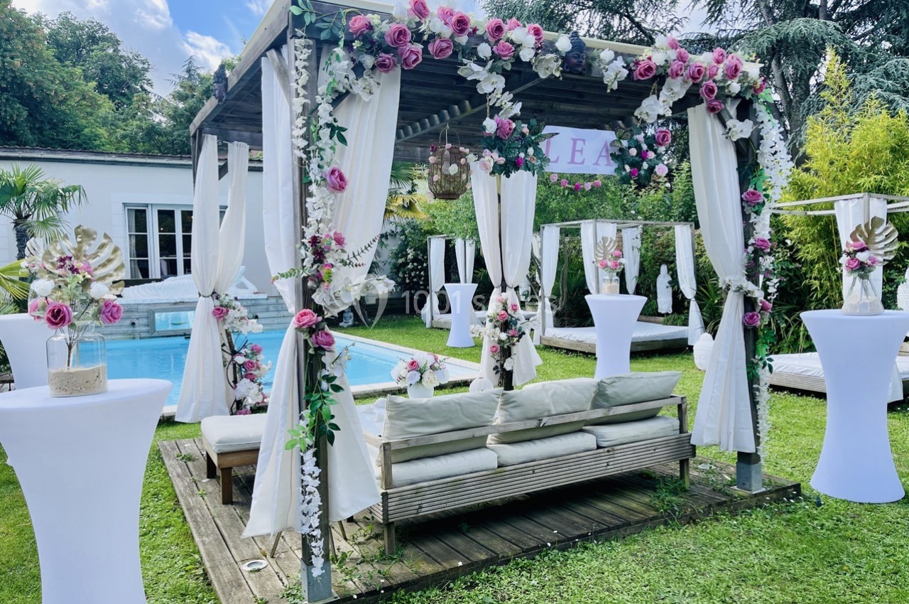Pergola décorée de fleurs et de voilages blancs près d'une piscine, entourée de mobilier de jardin et de verdure.