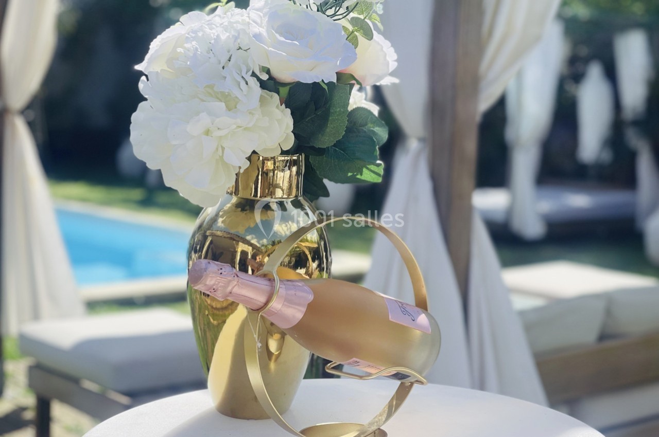 Vase doré avec des fleurs blanches et bouteille de champagne rosé dans un support, près d'une piscine et de lits extérieurs.