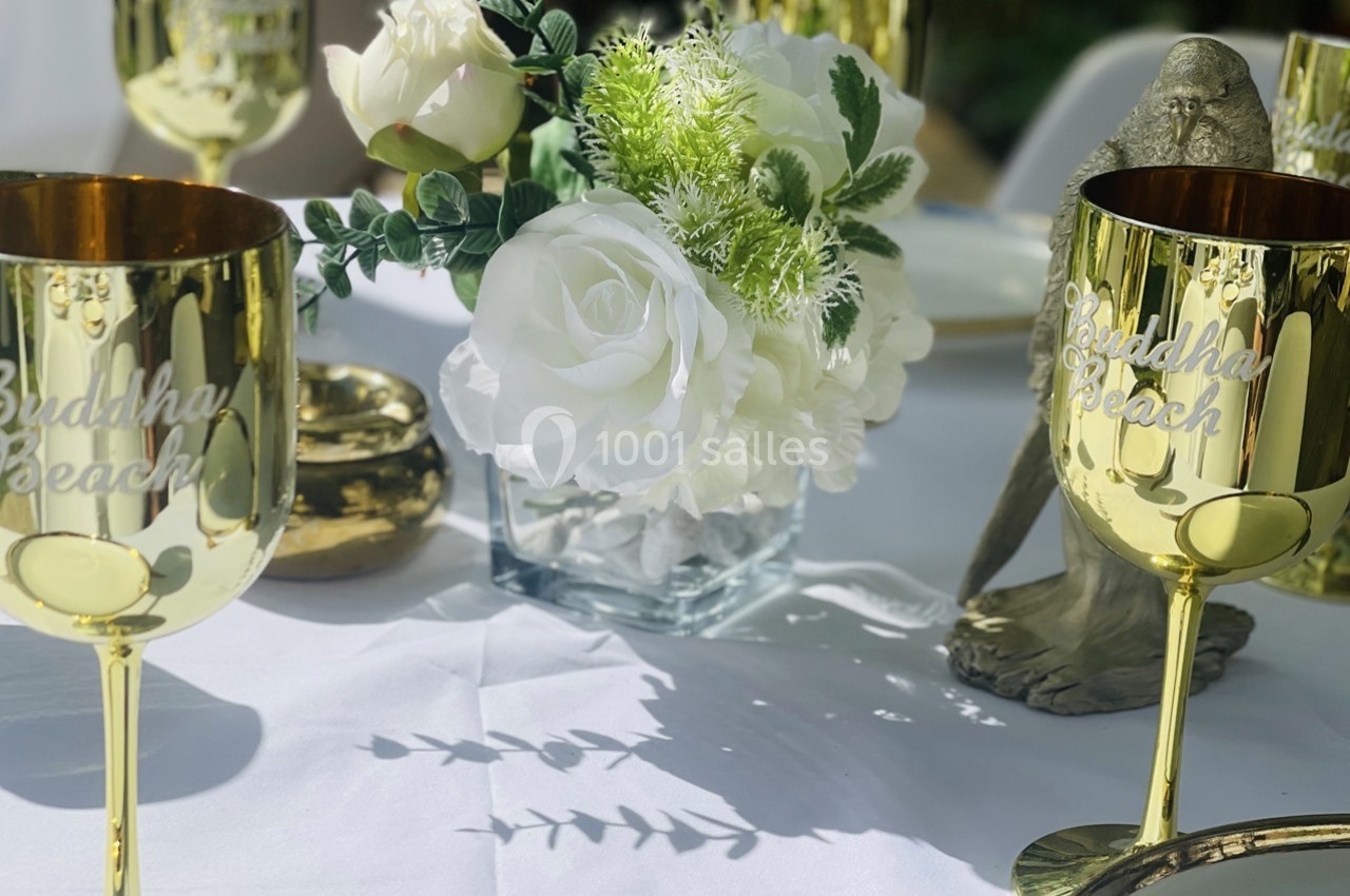 Centre de table avec roses blanches, verdure et verres dorés gravés ’Buddha Beach’ sur une nappe blanche.