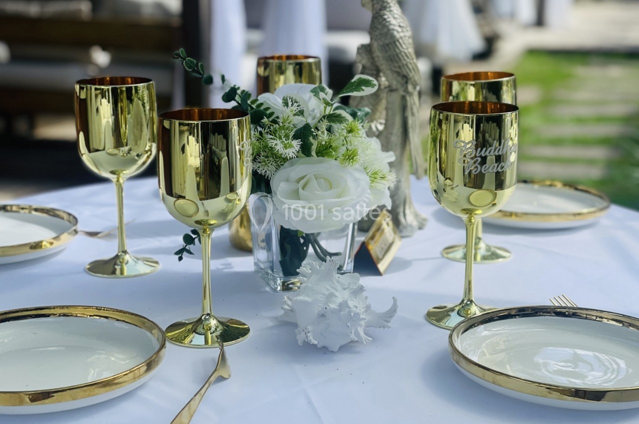Table dressée avec des assiettes blanches, couverts dorés, verres dorés et un bouquet de fleurs blanches au centre.