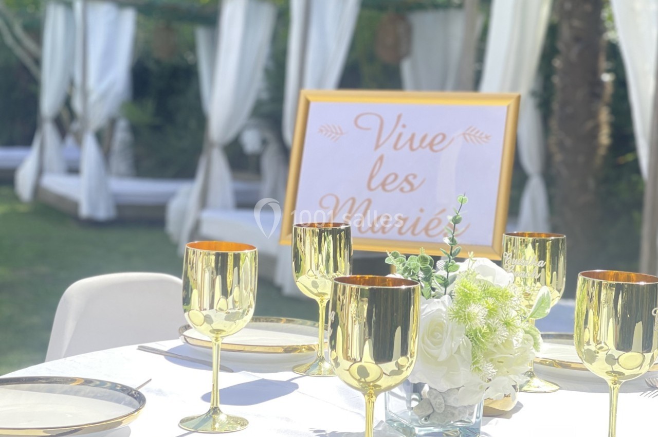 Table décorée avec des verres dorés et un centre de table floral, avec un panneau ’Vive les Mariés’ en arrière-plan.