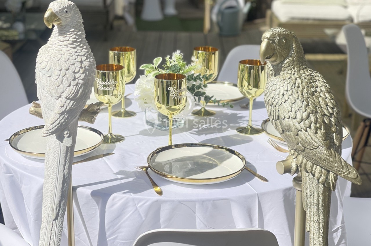 Deux statues de perroquets décorent une table dressée avec des verres dorés et une vaisselle élégante en extérieur.