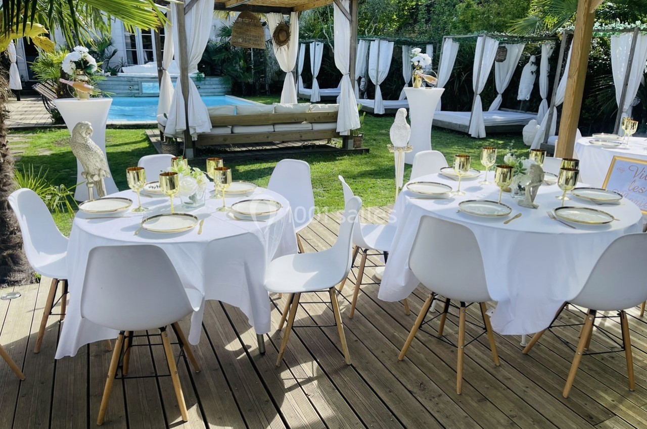 Tables dressées avec nappes blanches, vaisselle élégante et chaises modernes sur une terrasse en bois, jardin en arrière…
