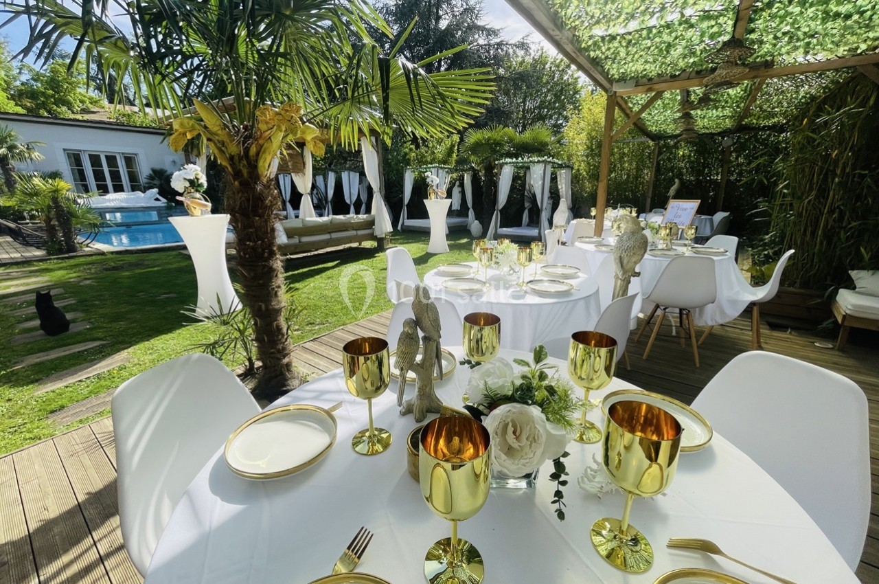 Table dressée avec vaisselle dorée sur une terrasse en bois, entourée de végétation luxuriante et d'une piscine en arrière…