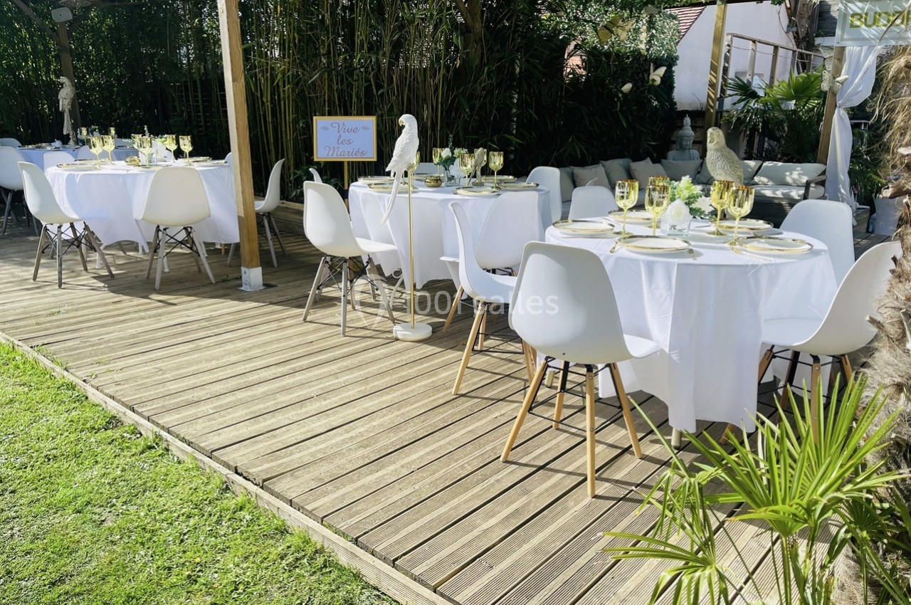 Tables rondes dressées avec nappes blanches et chaises modernes sur une terrasse en bois, entourée de verdure.