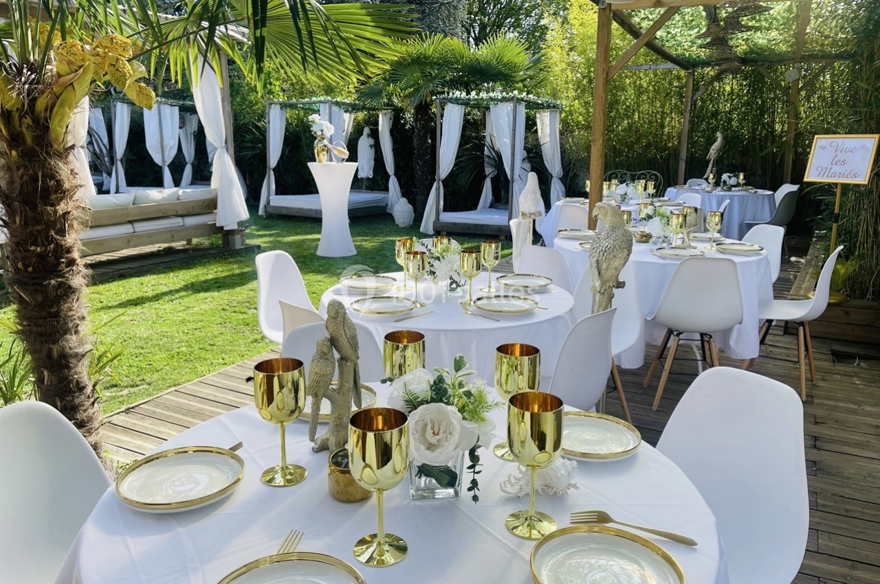 Tables élégamment dressées avec nappes blanches, vaisselle dorée et décoration florale, dans un jardin verdoyant.