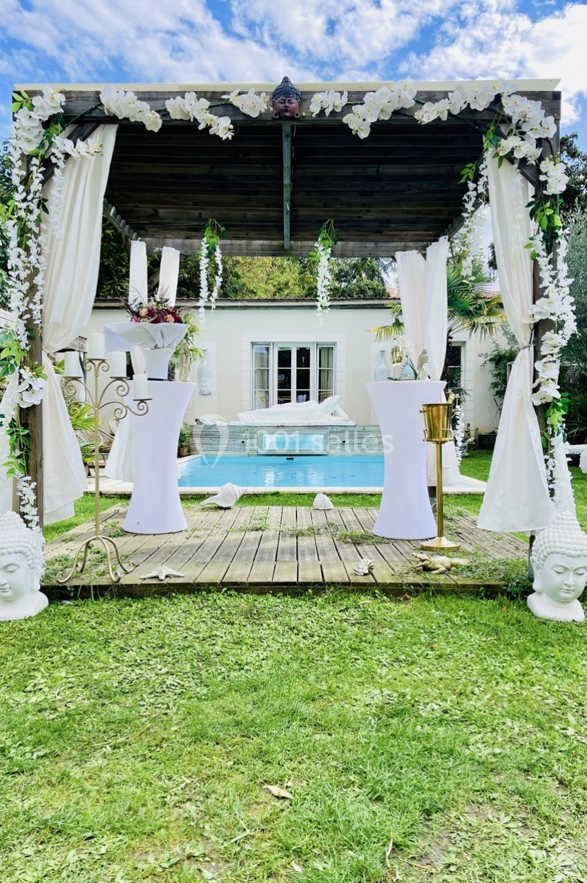 Tonnelle décorée de rideaux blancs et de fleurs, située devant une piscine dans un jardin verdoyant.
