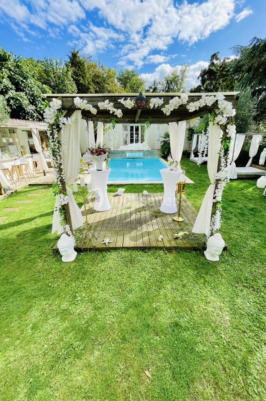 Pergola décorée de fleurs blanches devant une piscine, entourée de verdure et de mobilier extérieur élégant.