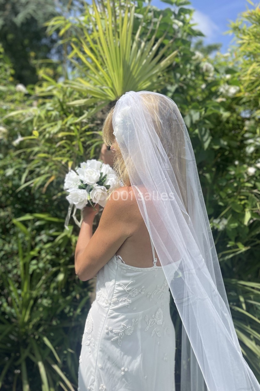 Une mariée en robe blanche et voile tient un bouquet de fleurs blanches devant un jardin verdoyant.
