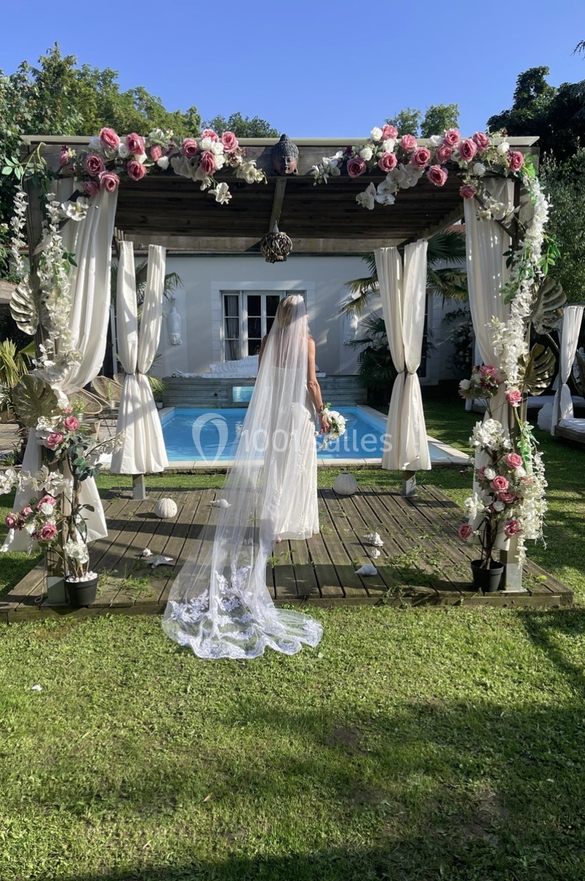 Une mariée en robe blanche et voile long se tient sous une arche fleurie devant une piscine dans un jardin.
