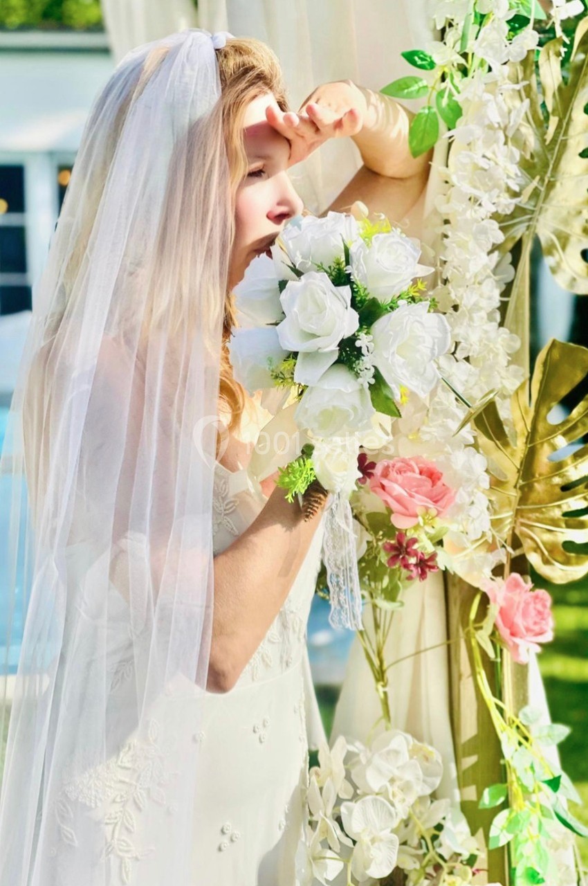Une mariée en robe blanche et voile tient un bouquet de fleurs blanches près d'une arche décorée de feuillages et fleurs.