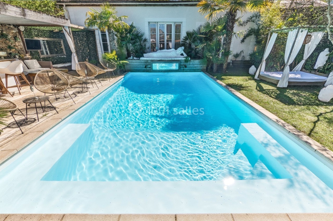 Piscine extérieure rectangulaire entourée de chaises, végétation et lits à baldaquin dans un jardin ensoleillé.