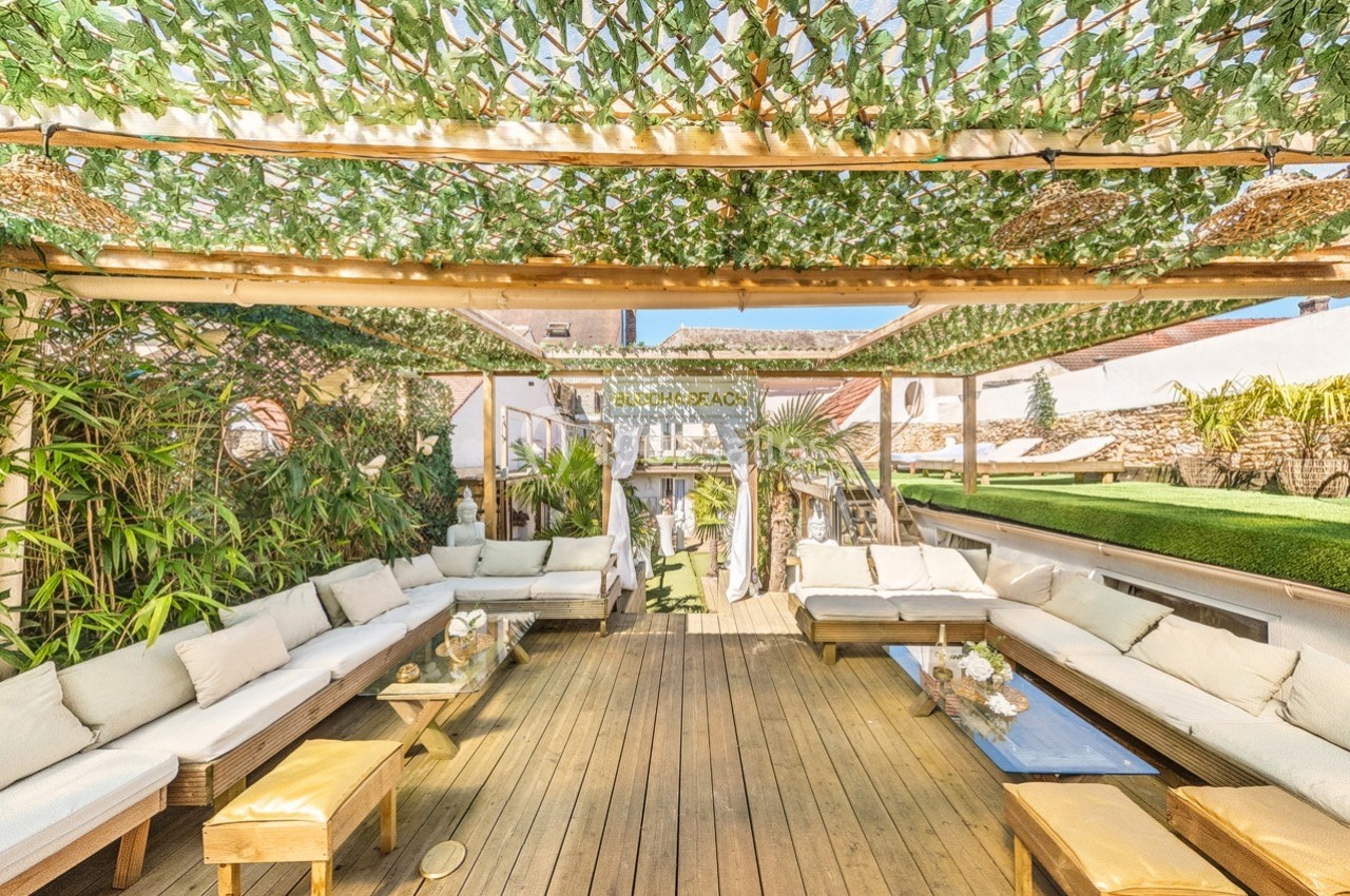 Terrasse en bois avec canapés, tables basses et pergola couverte de feuillage, entourée de verdure.
