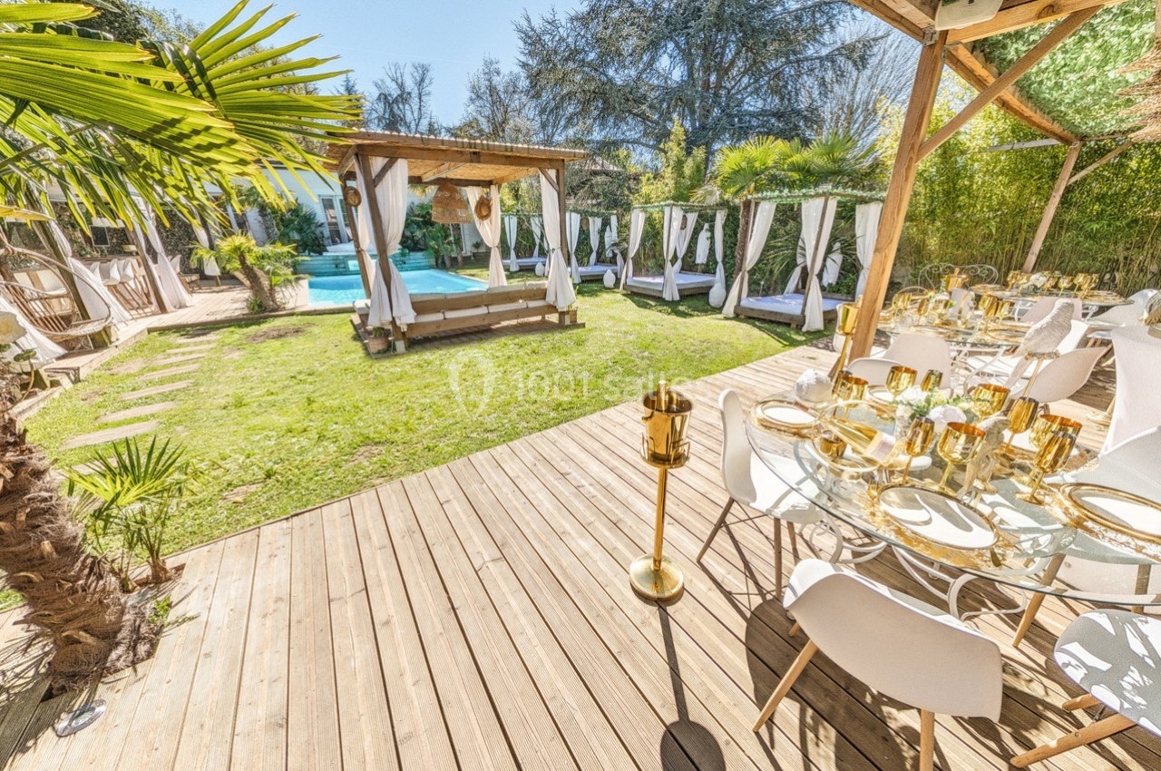 Terrasse en bois avec tables dressées, vue sur un jardin avec piscine, lits à baldaquin et végétation luxuriante.