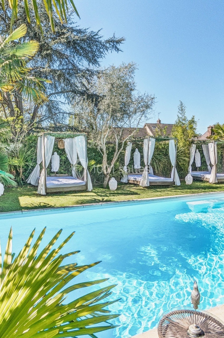 Piscine extérieure entourée de lits balinais avec rideaux blancs, végétation luxuriante et ciel dégagé.
