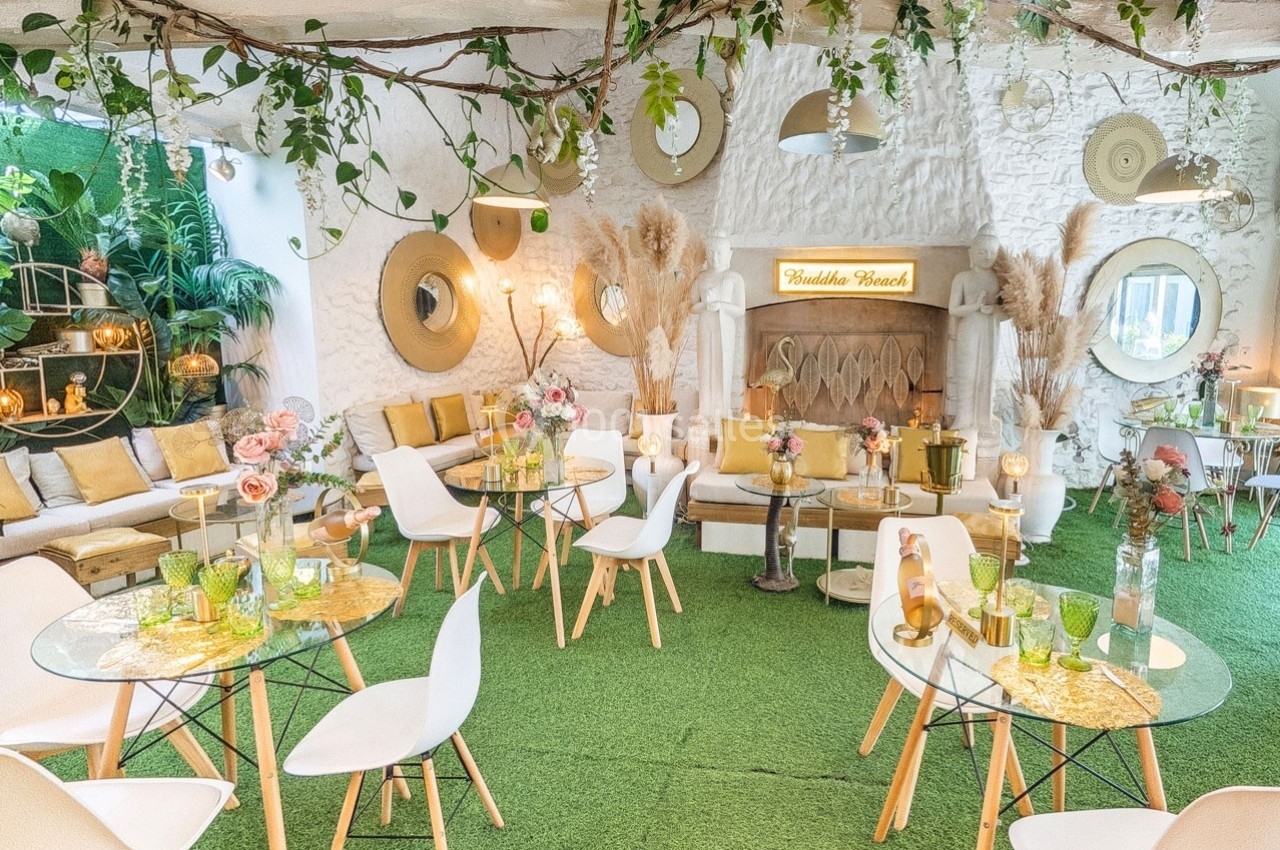 Salle lumineuse décorée de plantes, mobilier moderne et tables en verre sur tapis vert, ambiance conviviale et naturelle.