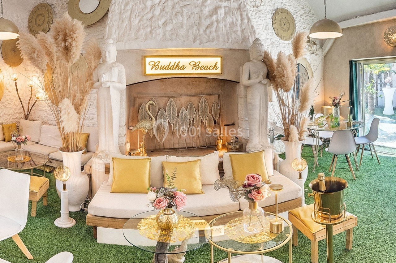 Salon lumineux avec décoration exotique, statues, végétation, coussins dorés et enseigne lumineuse ’Buddha Beach’.