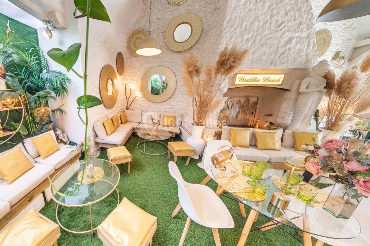 Salon lumineux avec décoration bohème, coussins dorés, miroirs ronds, plantes et tables en verre sur tapis vert.