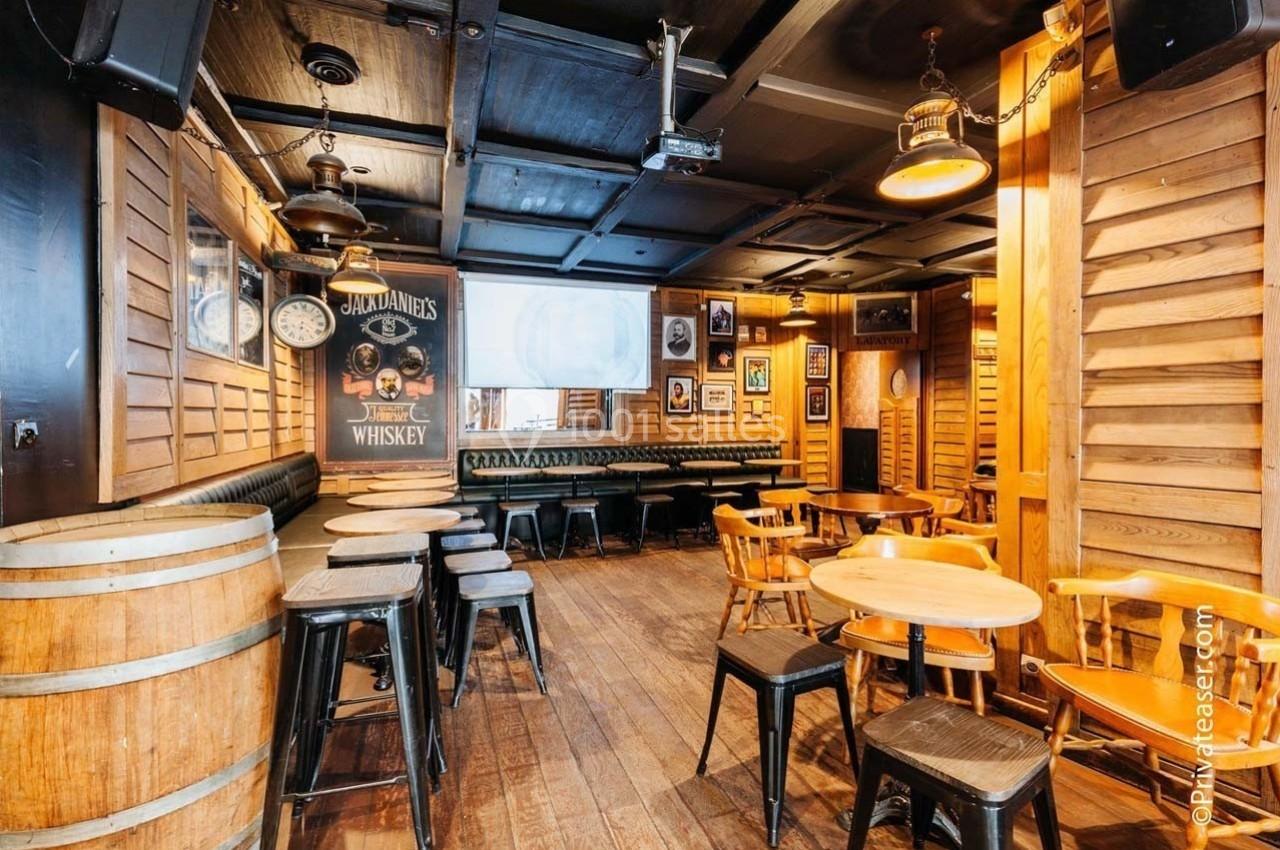 Salle de bar en bois avec tables, chaises, tonneaux décoratifs et affiches murales, éclairée par des lampes suspendues.