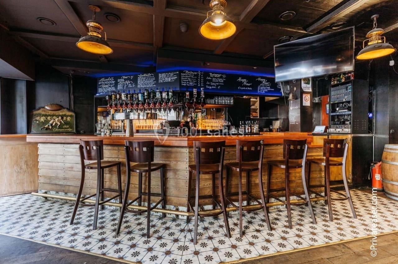 Bar en bois avec tabourets alignés, étagères de bouteilles et ardoises affichant des menus, éclairage chaleureux.