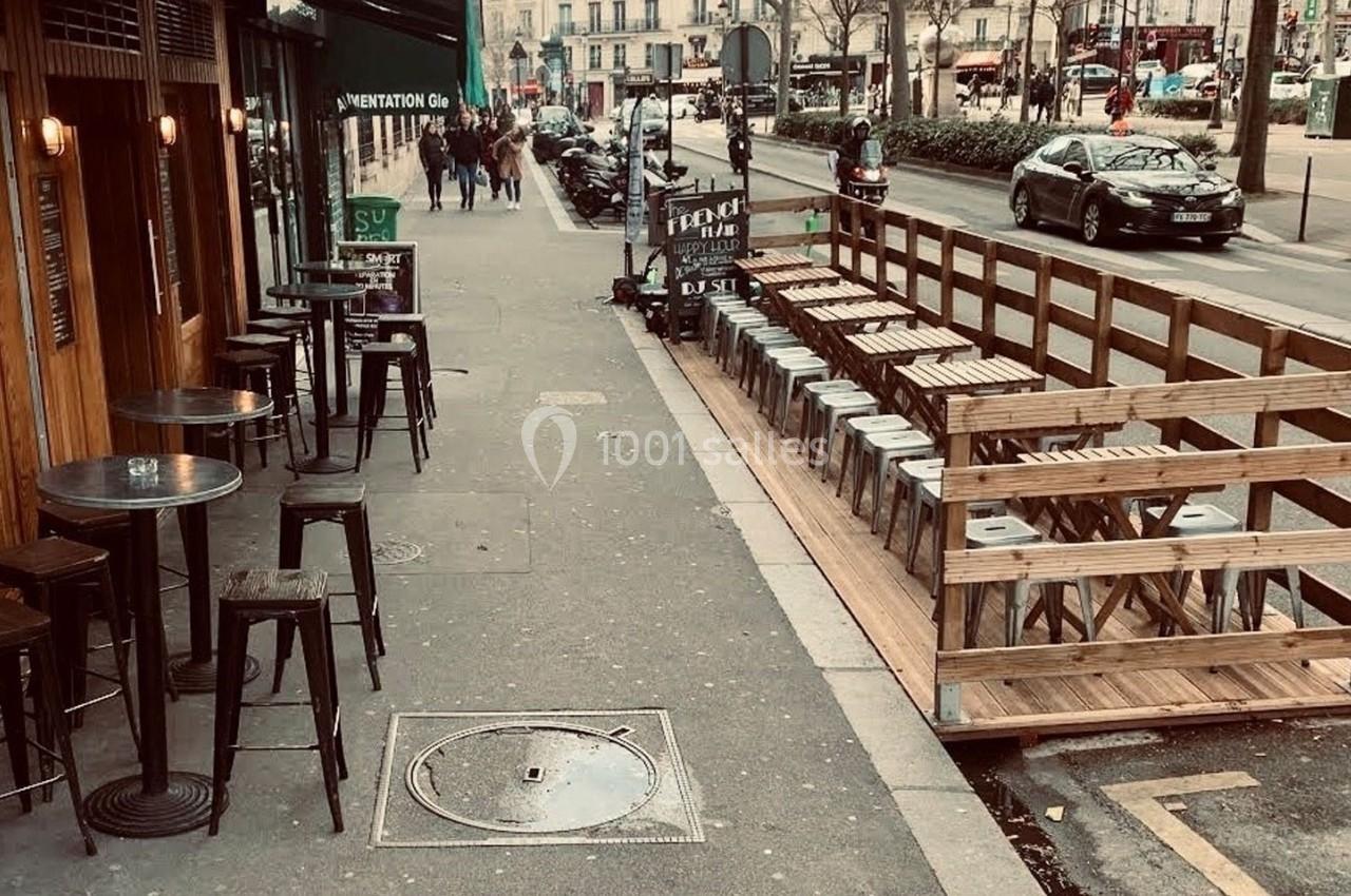 Terrasse en bois avec tabourets alignés sur un trottoir, à côté d'un café et d'une rue passante en ville.