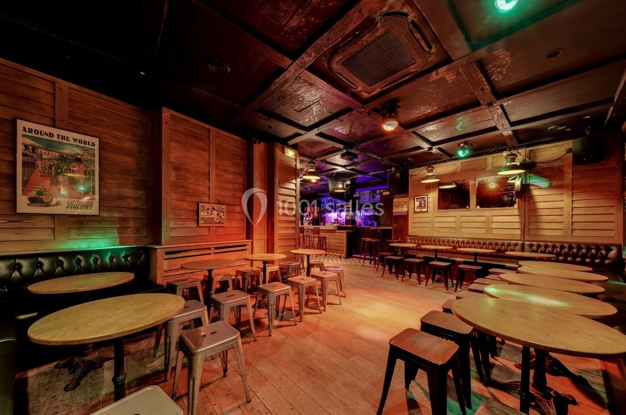 Salle de bar avec tables rondes en bois, tabourets métalliques et éclairage tamisé dans une ambiance chaleureuse.