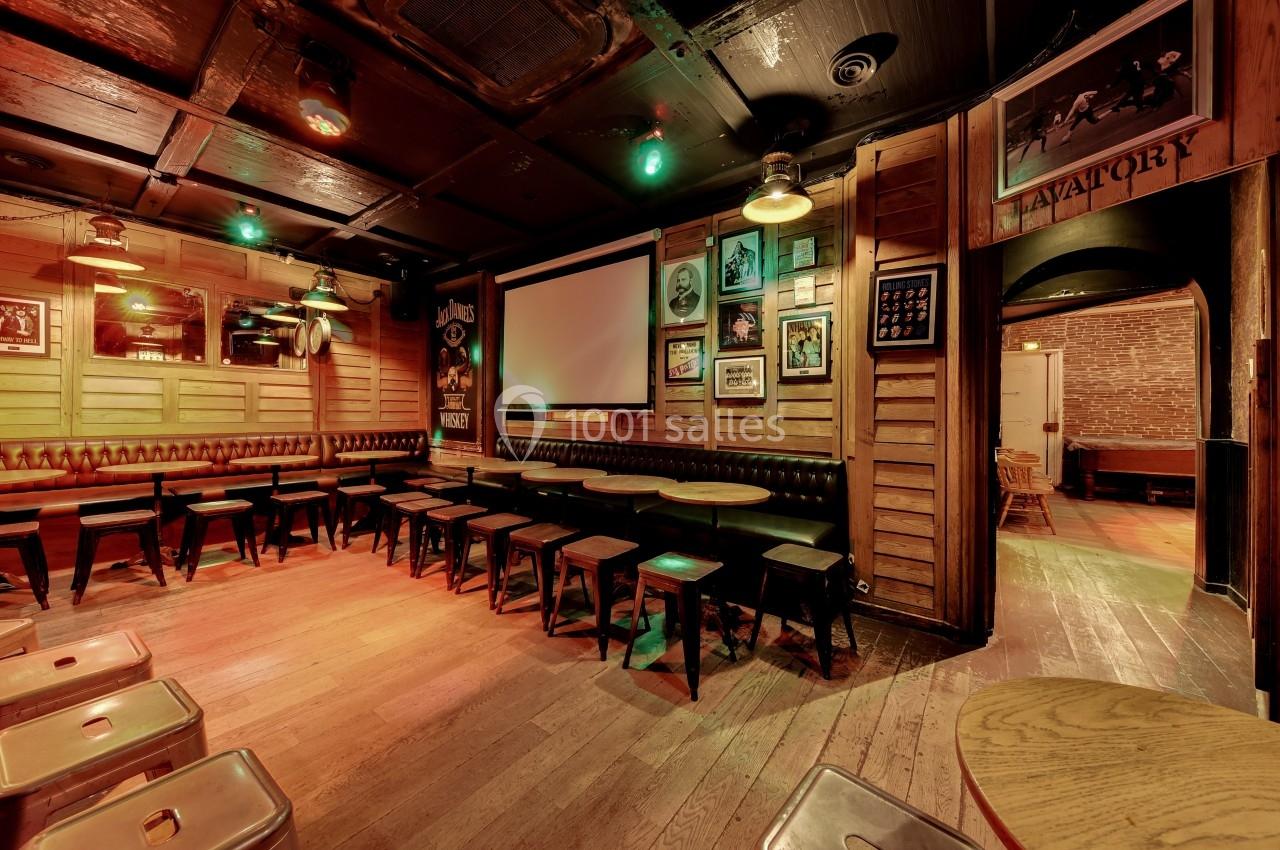 Salle de bar chaleureuse avec tables en bois, tabourets alignés, écran mural et décorations rétro sur les murs.