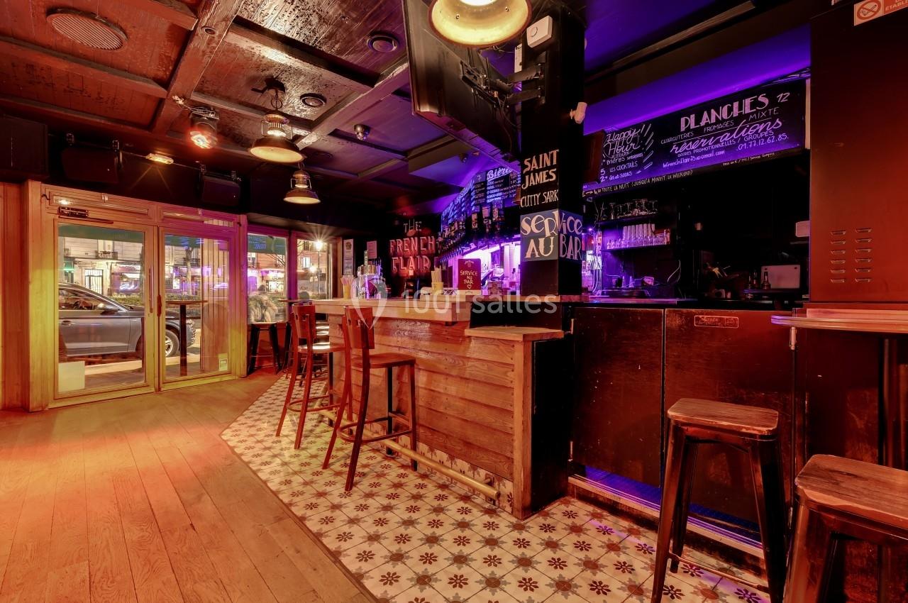 Intérieur d'un bar avec comptoir en bois, tabourets hauts, éclairage chaleureux et tableau noir avec menu écrit à la craie.