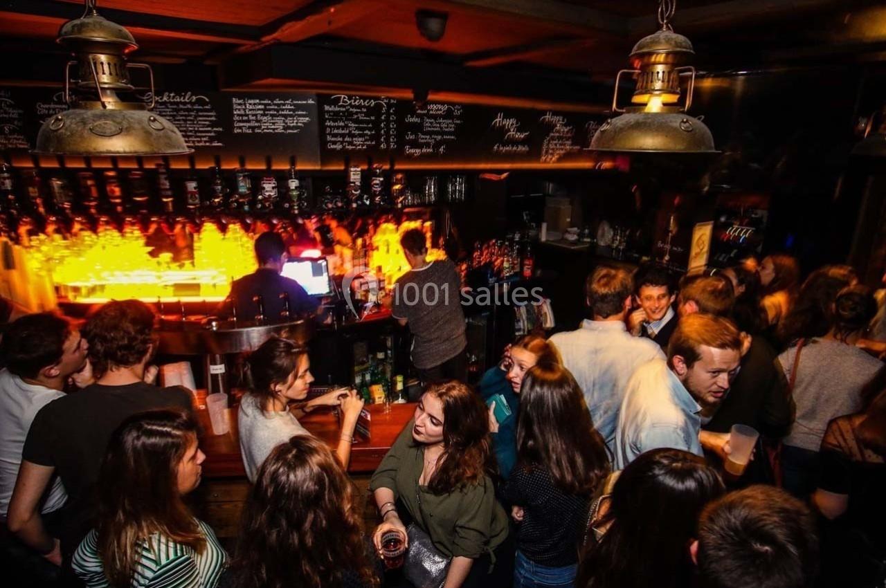 Des personnes discutent et boivent dans un bar animé, éclairé par des lumières chaudes près du comptoir.