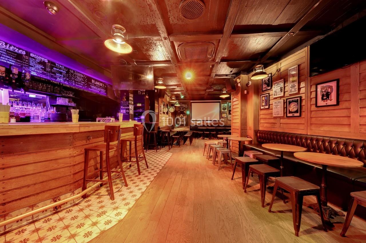 Intérieur d'un bar avec comptoir en bois, tabourets, tables basses, éclairage tamisé et écran au fond.
