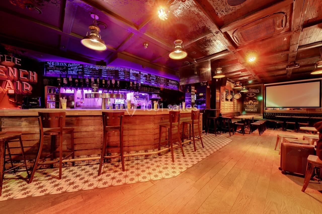 Intérieur d'un bar avec un long comptoir en bois, des tabourets hauts et un éclairage tamisé.