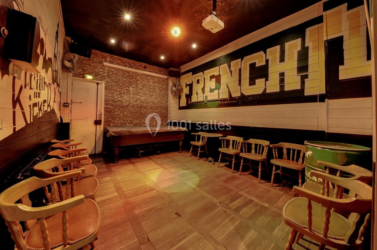 Salle vide avec des chaises en bois disposées autour de tables, murs en briques et inscription ’FRENCH’ sur un mur.