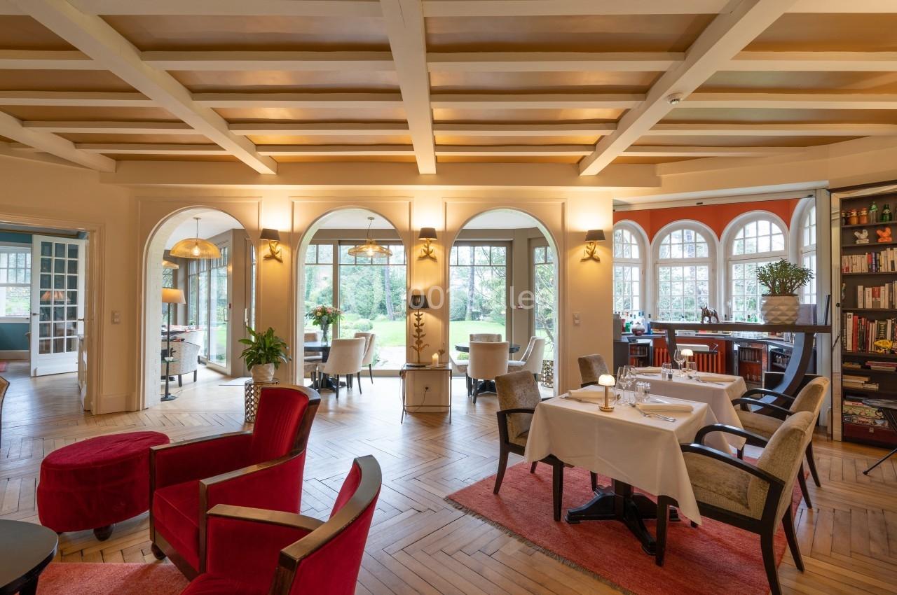 Salle lumineuse avec tables dressées, fauteuils rouges et vue sur un jardin à travers de grandes fenêtres cintrées.