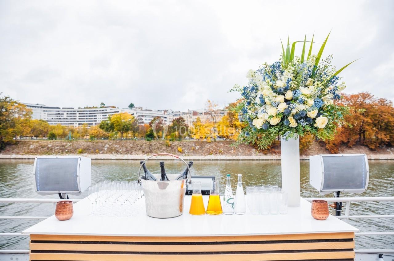 Table dressée en extérieur avec seau à champagne, verres, carafes de jus et bouquet floral, au bord d'une rivière.