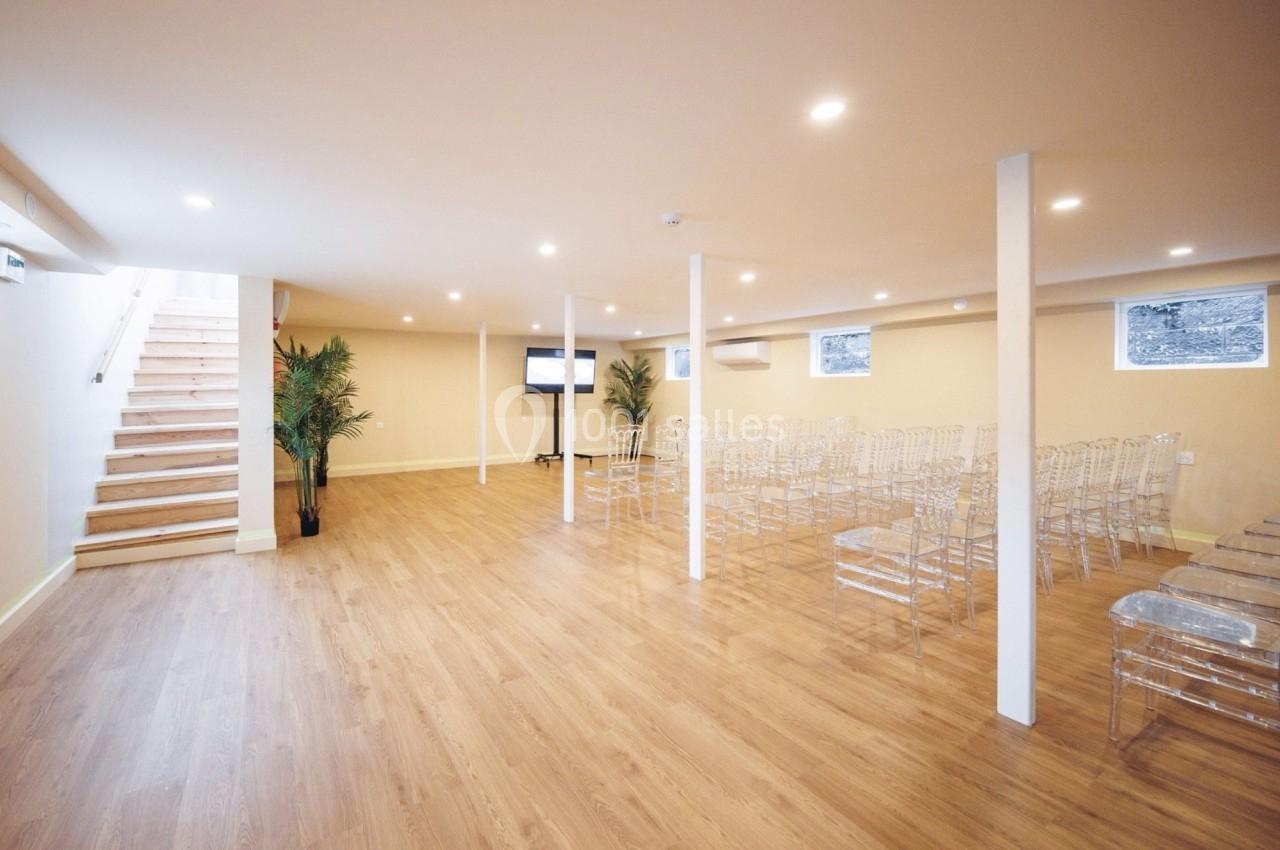 Salle lumineuse avec parquet, chaises transparentes alignées, écran au fond et escalier menant à l'étage.