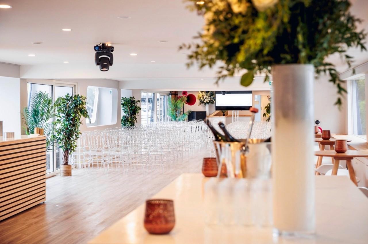 Salle lumineuse aménagée pour un événement, avec des chaises transparentes, des plantes et un bar au premier plan.