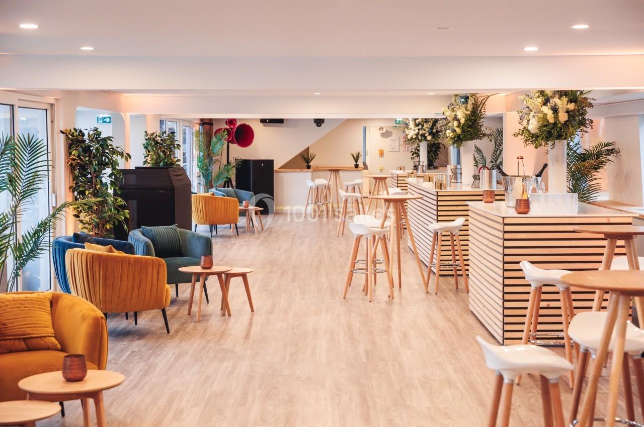 Salle lumineuse avec mobilier moderne, tabourets hauts, fauteuils colorés, plantes et comptoirs en bois clair.