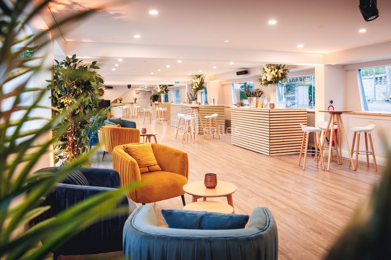 Salle lumineuse avec fauteuils colorés, tables basses, bar en bois clair et plantes décoratives.