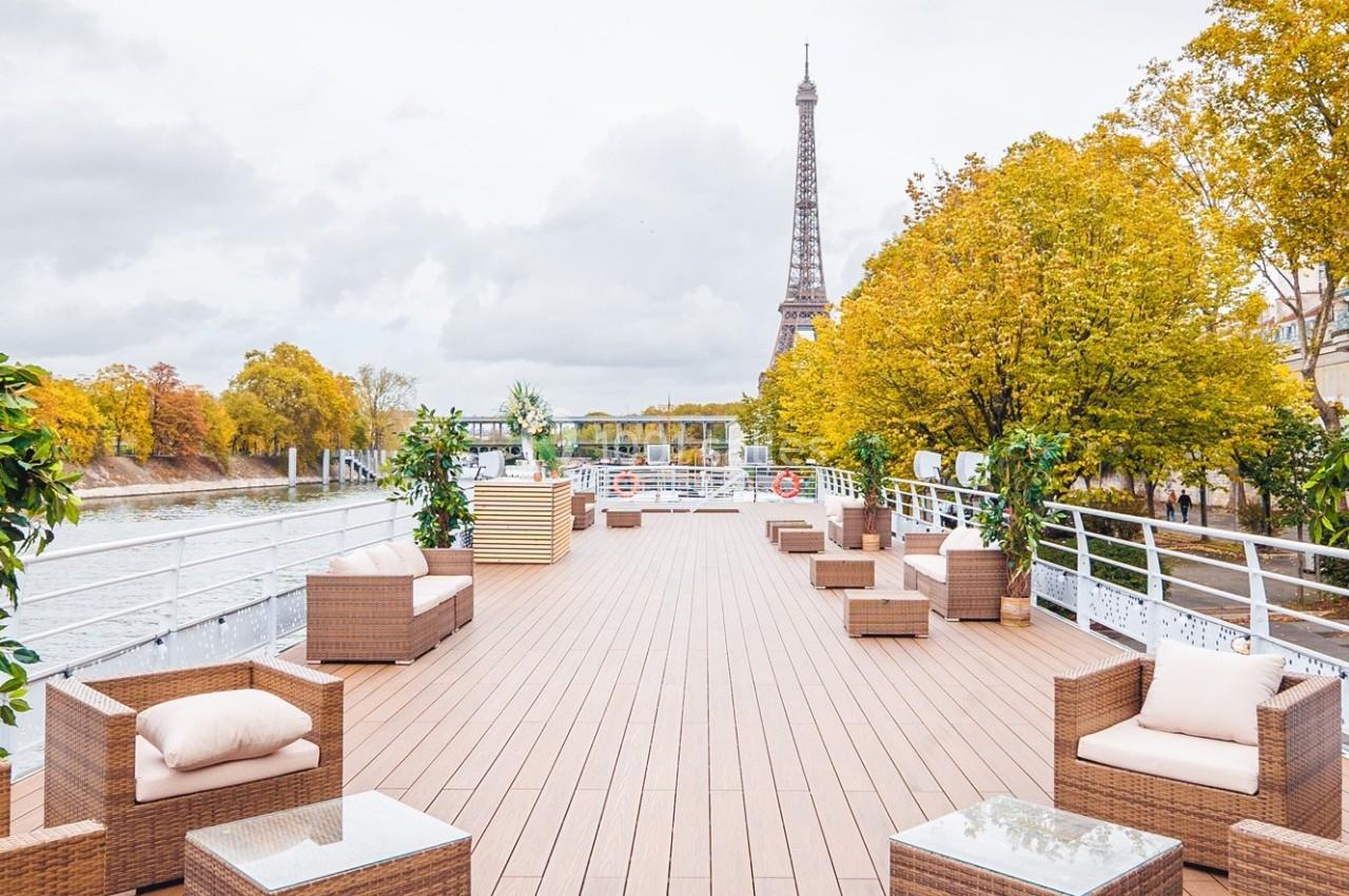 Terrasse en bois aménagée avec des fauteuils et tables basses, vue sur la Seine et la tour Eiffel en arrière-plan.