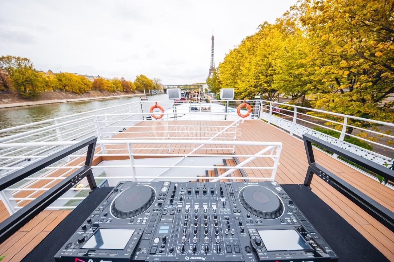 Console DJ sur un bateau avec vue sur la Seine, des arbres aux couleurs automnales et la tour Eiffel en arrière-plan.