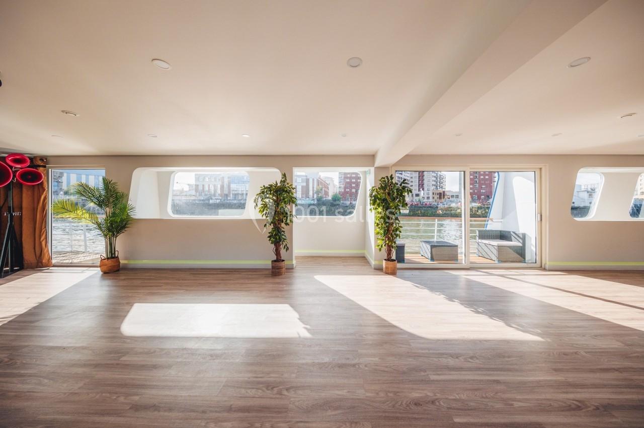Salle lumineuse avec parquet, grandes fenêtres donnant sur l'extérieur, plantes en pot et vue sur des bâtiments.